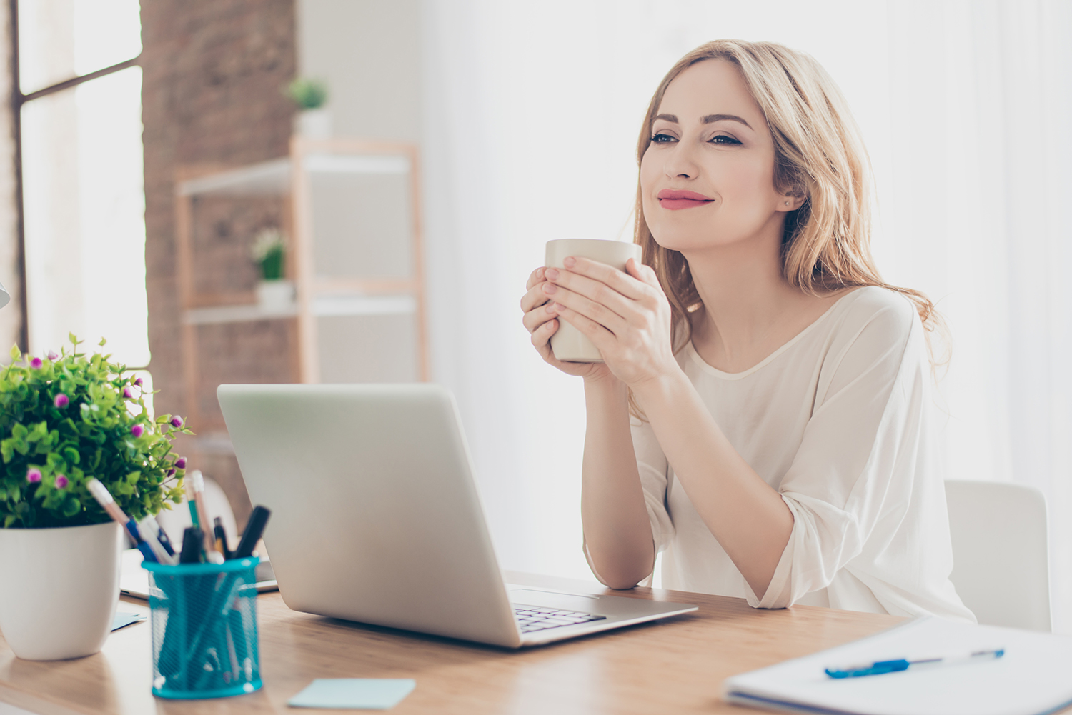 A content woman at her desk, in front of her laptop, and enjoying a hot beverage